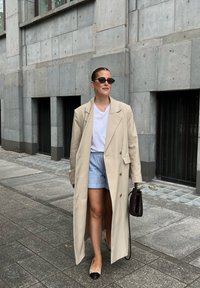 Casaco trench bege, t-shirt branca, calções listrados azuis, sapatilhas pretas e mala preta fina. Óculos de sol escuros e cabelo liso. Fundo urbano.