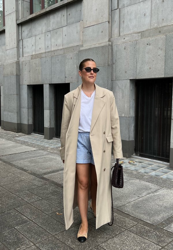 Trenci bej, tricou alb, pantaloni scurți albaştri cu dungi, balerini negri și geantă neagră subțire. Ochelari de soare întunecați și păr neted. Fundal urban.