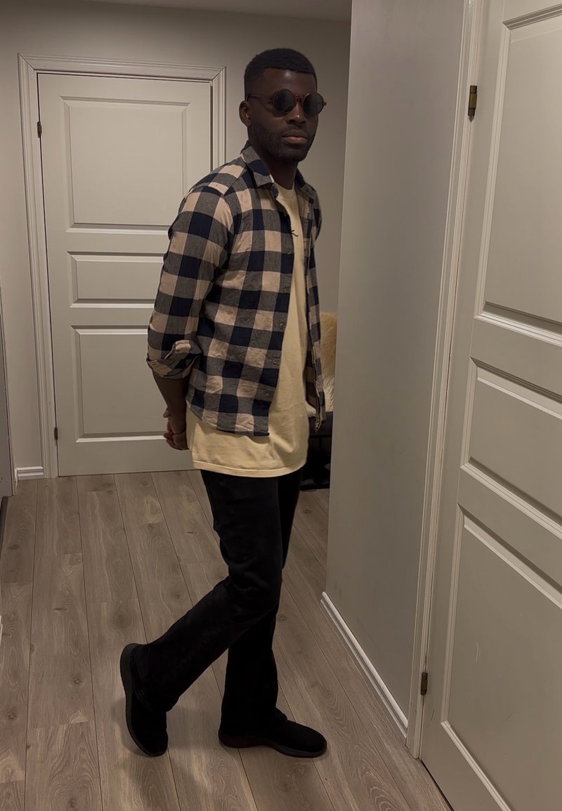 Homme portant des lunettes de soleil, une chemise beige avec un surchemise à carreaux bleu marine, et un pantalon noir, debout à l'intérieur près de portes blanches sur un sol en bois clair.