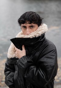 Persoon met nat krullend haar die een zwarte leren jas met crème kleurige schapenvachtkraag omhoog trekt, staand bij een vage achtergrond van een waterlichaam.