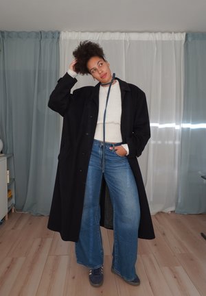 Abrigo largo negro, suéter blanco con textura, vaqueros azules de cintura alta y zapatillas azules, en una habitación luminosa con cortinas translúcidas.