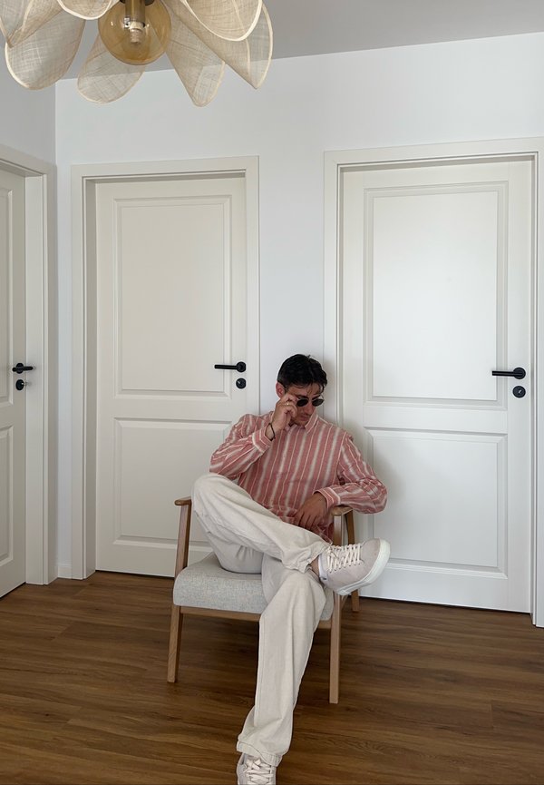 Uomo che indossa una camicia rosa a righe e occhiali da sole, seduto a gambe incrociate su una sedia davanti a tre porte bianche chiuse.