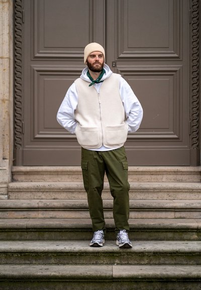 Chaleco de forro polar beige sobre una sudadera blanca, bandana verde, pantalones cargo verdes y zapatillas grises. De pie en los escalones frente a una puerta marrón.