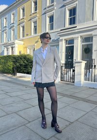 Blazer beige oversize, chemise blanche, jupe noire à fines rayures, collants noirs transparents, et mocassins à talons hauts violets dans un cadre urbain.