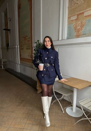 Mujer con traje de falda azul marino y botas blancas sosteniendo una taza de café, de pie junto a una mesa y sillas en el interior con murales decorativos en la pared.