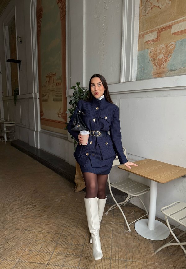 Mujer con traje de falda azul marino y botas blancas sosteniendo una taza de café, de pie junto a una mesa y sillas en el interior con murales decorativos en la pared.