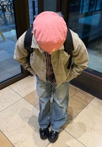 Persoană îmbrăcată cu o pălărie roz, cămașă cu carouri, jachetă din catifea și blugi lejeri albaștri. Pantofi negri strălucitori. Poziție de a se pleca într-un cadru interior.