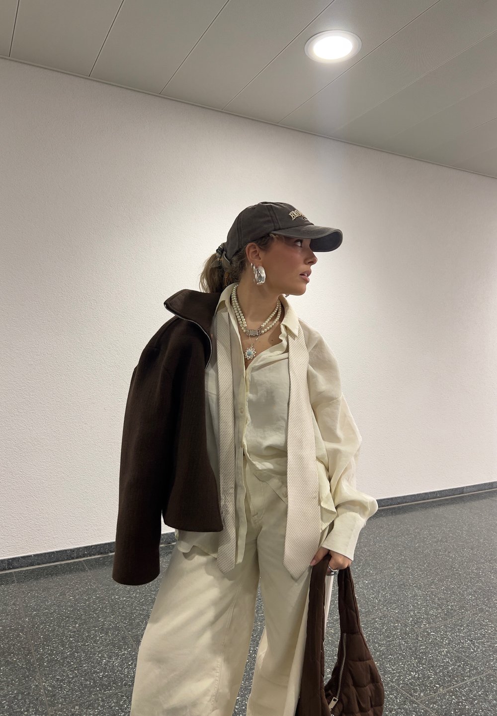 Ljust färgad, oversize outfit med en texturerad beige skjorta och vida byxor. Drapering av en mörkbrun jacka och ett vadderat brunt väsk. Tillbehör inkluderar en keps och flerlagrade halsband.