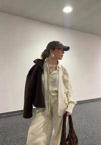 Ljust färgad, oversize outfit med en texturerad beige skjorta och vida byxor. Drapering av en mörkbrun jacka och ett vadderat brunt väsk. Tillbehör inkluderar en keps och flerlagrade halsband.