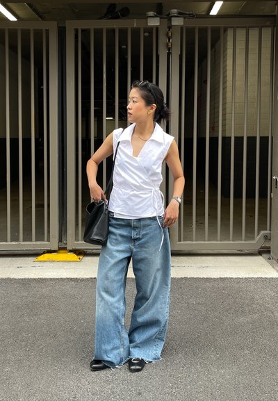 Camisa blanca sin mangas con un cuello, combinada con jeans de pierna ancha de color azul claro. Un bolso negro y sandalias completan el conjunto.