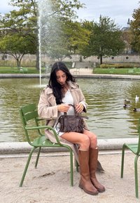 Donna con trench beige e stivali marroni seduta su una sedia verde vicino a una fontana, con in mano una borsa marrone scuro, circondata da alberi e anatre.