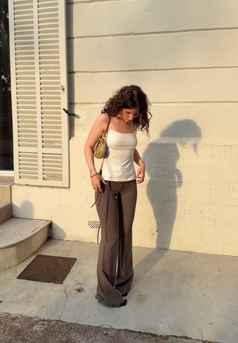 Femme aux cheveux bouclés regarde vers le bas en se tenant près d'un mur beige avec des volets fermés, portant un haut blanc, un pantalon marron et tenant un petit sac.