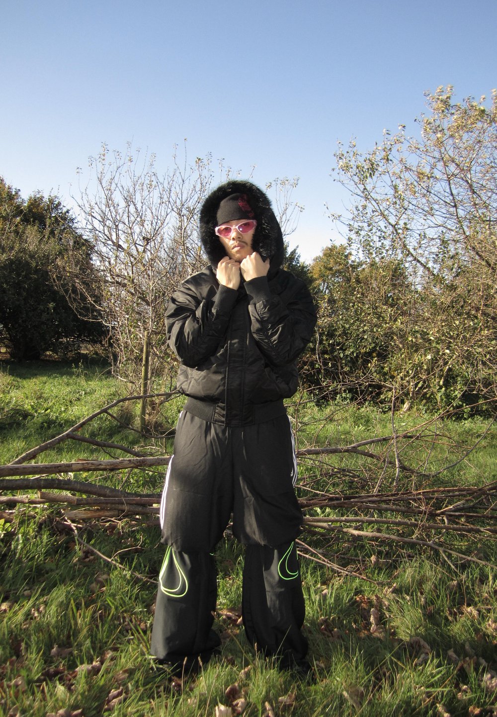 Casaco preto com capuz forrado de pele, calças pretas com detalhes em verde néon e óculos de sol cor-de-rosa. Em pé numa área com relva e árvores ao fundo.