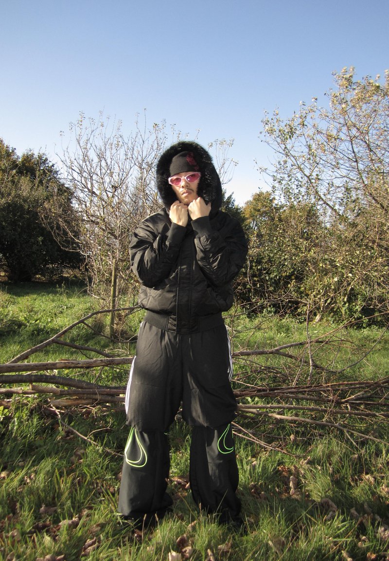 Giacca nera con cappuccio foderato in pelliccia, pantaloni neri con dettagli verde neon e occhiali da sole rosa. In piedi in un'area erbosa con alberi sullo sfondo.