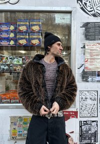 Brown faux fur jacket with black leather accents, zippered front, worn over a patterned grey shirt, paired with black trousers and a beanie.