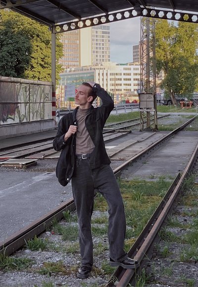 Figura que lleva una chaqueta negra abotonada sobre un suéter de cuello alto beige, jeans negros y botas, de pie sobre las vías del tren con vegetación.