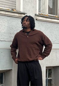 Brązowa, teksturowana koszulka polo z długim rękawem, kołnierzykiem i dwoma guzikami, w połączeniu z czarnymi plisowanymi spodniami. Prosty design, swobodny krój.