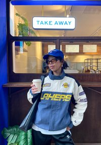 Veste à thème Lakers en blanc et bleu marine avec logo jaune, associée à un pantalon noir. Tenant une tasse de café, debout devant un comptoir de café.