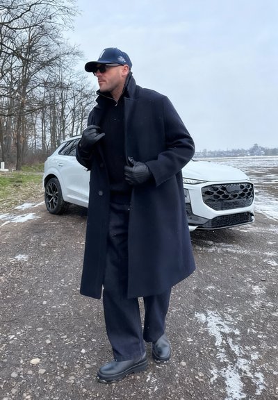 Hombre con abrigo negro, guantes, gorra y gafas de sol de pie en una carretera nevada junto a un SUV blanco, con un bosque y un campo al fondo.