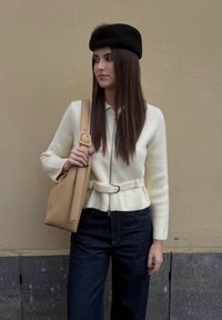Femme aux longs cheveux foncés portant un béret noir, un pull blanc zippé avec ceinture, un jean sombre, tenant un sac à bandoulière beige devant un mur couleur sable.