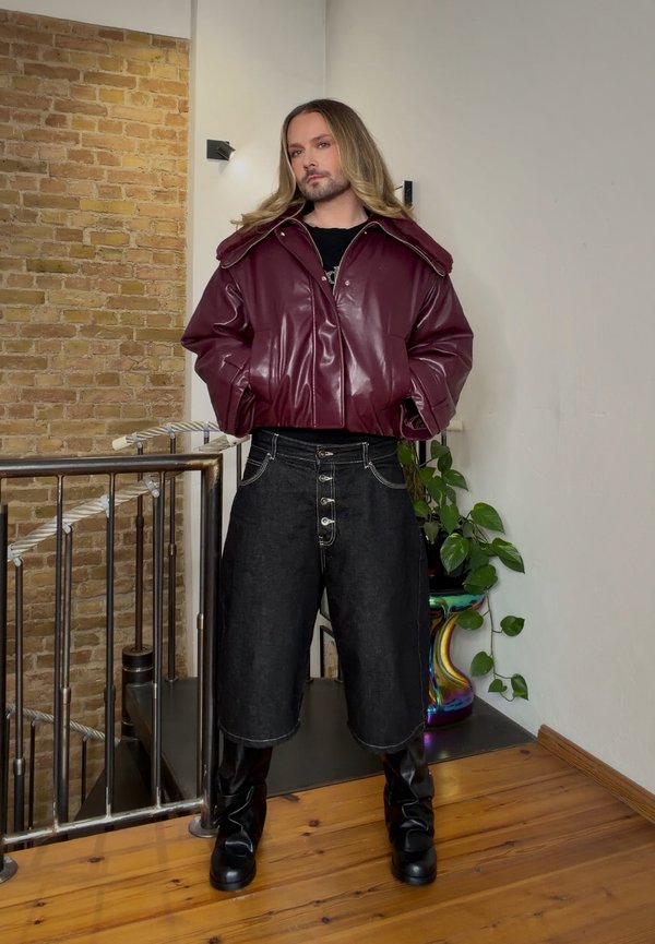Una persona posa en un interior con una chaqueta de cuero burdeos, pantalones cortos negros hasta la rodilla y botas altas negras.