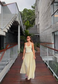 Lysegul haltertop og flydende maxinederdel lavet af letvægtsstof, båret med pink sandaler, på en trægangsti i en arkitektonisk setting.