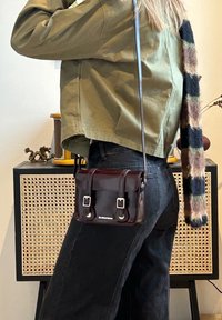 Personne portant une veste verte et un jean noir avec un sac bandoulière marron foncé et une longue écharpe rayée à l'intérieur près d'une armoire tissée.