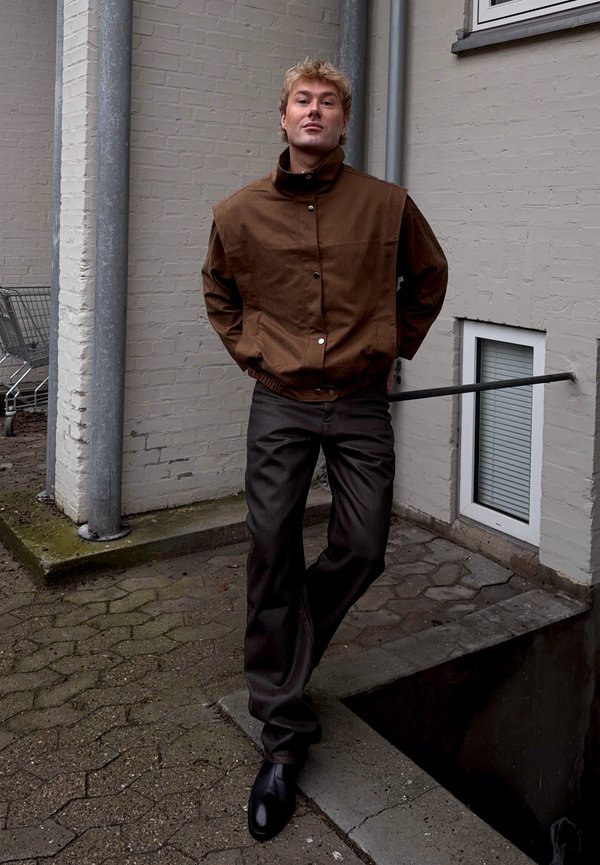 Jeune homme aux cheveux blonds courts portant une veste marron et un pantalon foncé, debout à l'extérieur contre un mur de briques clair près d'un chariot de supermarché.