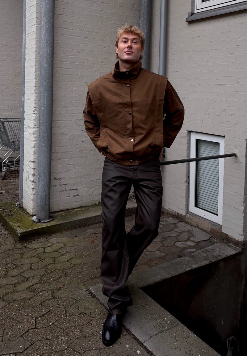 Jeune homme aux cheveux blonds courts portant une veste marron et un pantalon foncé, debout à l'extérieur contre un mur de briques claires près d'un chariot de supermarché.