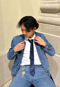 Jeans jakna preko bele srajce in mornarske kravate, sedeča, prilagaja jakno. Modri džins z srebrnimi ključi, gladka tekstura, naravna osvetlitev.