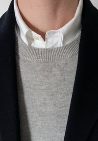 Gros plan d'une personne portant une chemise blanche boutonnée sous un pull en maille gris clair et une veste foncée.