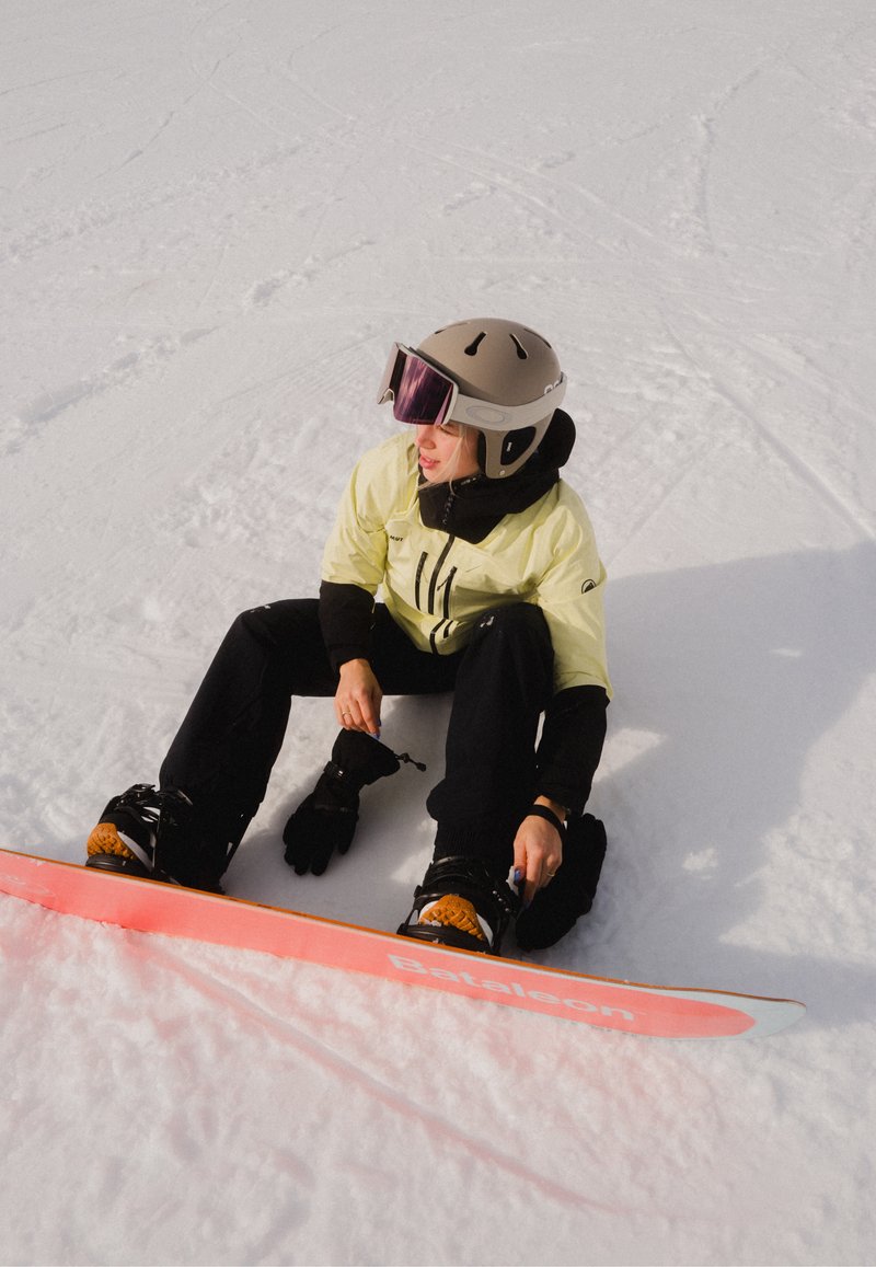 Personne portant un casque et des lunettes de protection attachant les fixations d'un snowboard en étant assise sur la neige en plein jour.