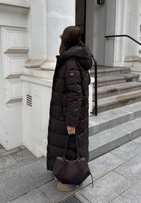 Mustade puffermantel kapuutsiga, millel on tikandmustrit, külgedel taskud ja logo. Kombineeritud tumeda pruuni käekoti ja beeži saapaga.