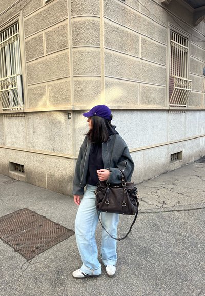 Una chaqueta bomber gris sobre una sudadera negra, jeans anchos de color azul claro, una gorra morada y un bolso negro con accesorios dorados. Ambiente urbano.
