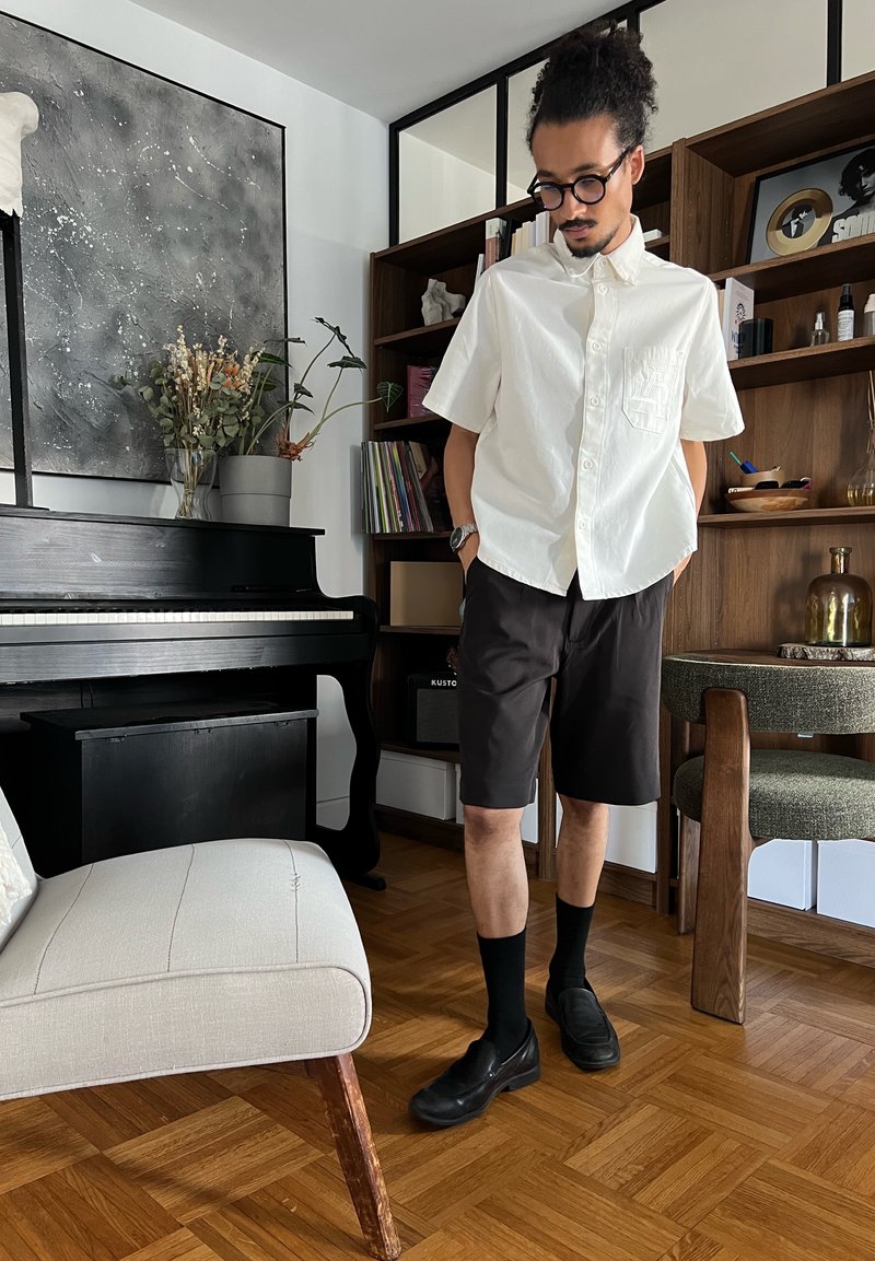 Homme aux cheveux bouclés et lunettes, portant une chemise blanche et un short noir, se tient à côté d'un piano dans un salon moderne avec des étagères en bois.