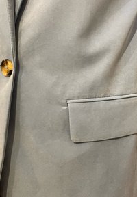Ανοιχτό γκρι σακάκι με λεία υφή υφάσματος, μία τσέπη στο στήθος και ξύλινο κουμπί στρογγυλής μορφής στη αριστερή πλευρά.