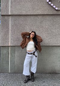 Hnedá kožená bomber bunda cez biely krátky top, svetlošedé voľné nohavice a tmavohnedé robustné topánky s prackou.