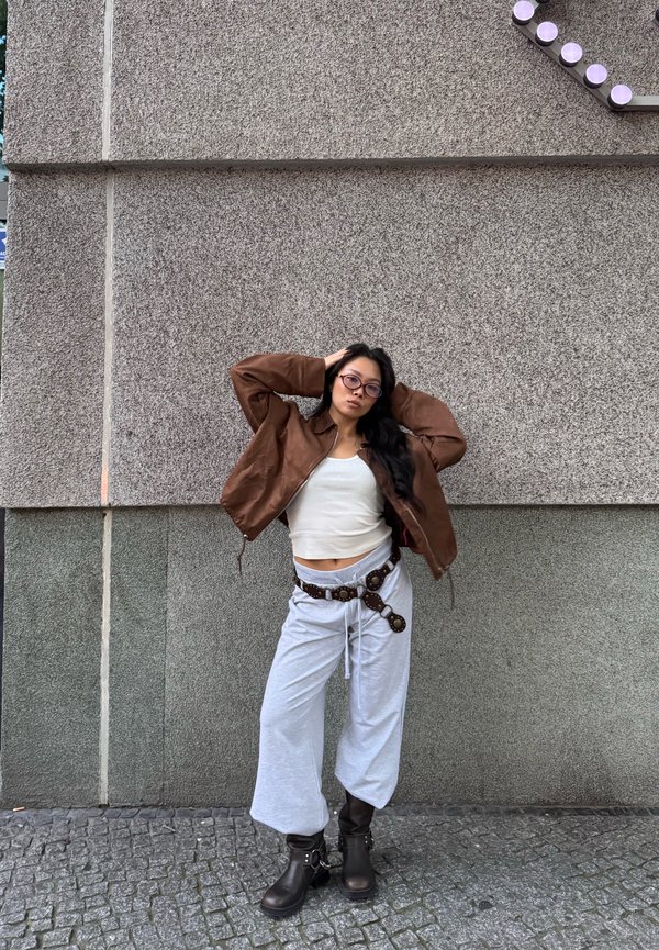 Brown leather bomber jacket over white cropped top, light gray wide-leg pants, and dark brown chunky boots with buckle detail.