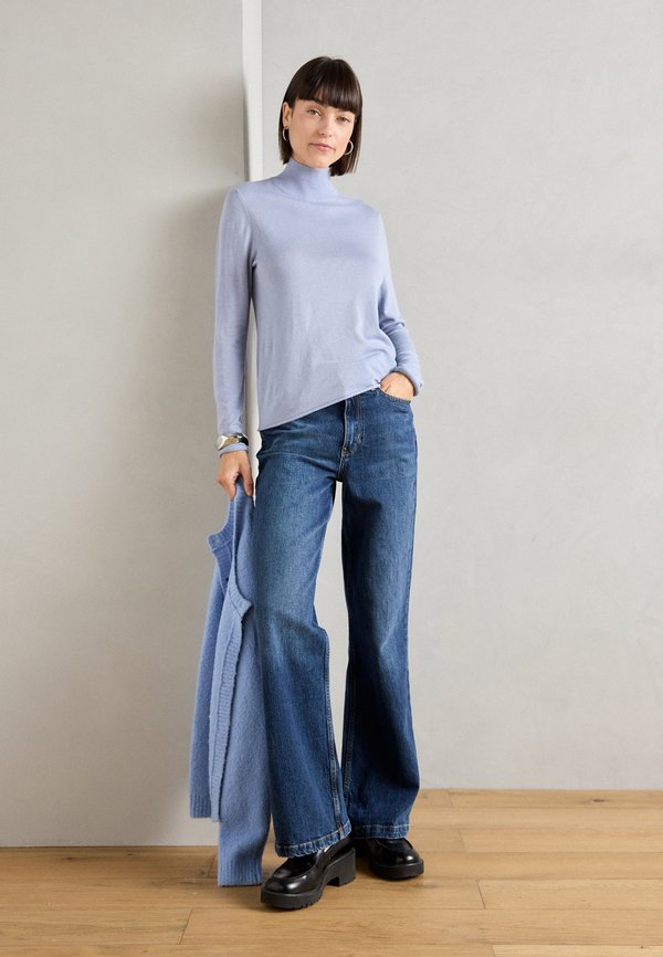 Mujer con suéter de cuello alto azul claro, jeans de pernera ancha, zapatos de plataforma negros, sosteniendo un cárdigan azul a juego, de pie sobre un suelo de madera.