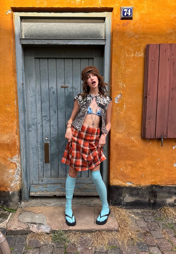 Jeune femme portant un haut de bikini bleu, un gilet argenté, une jupe à carreaux orange, des chaussettes bleues jusqu'aux genoux et des tongs, debout devant une porte bleue sur un mur orange.