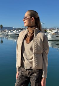 Femme portant des lunettes de soleil et une veste en tricot beige clair, debout près d'une marina avec des bateaux, portant une écharpe marron à motifs et un jean foncé.