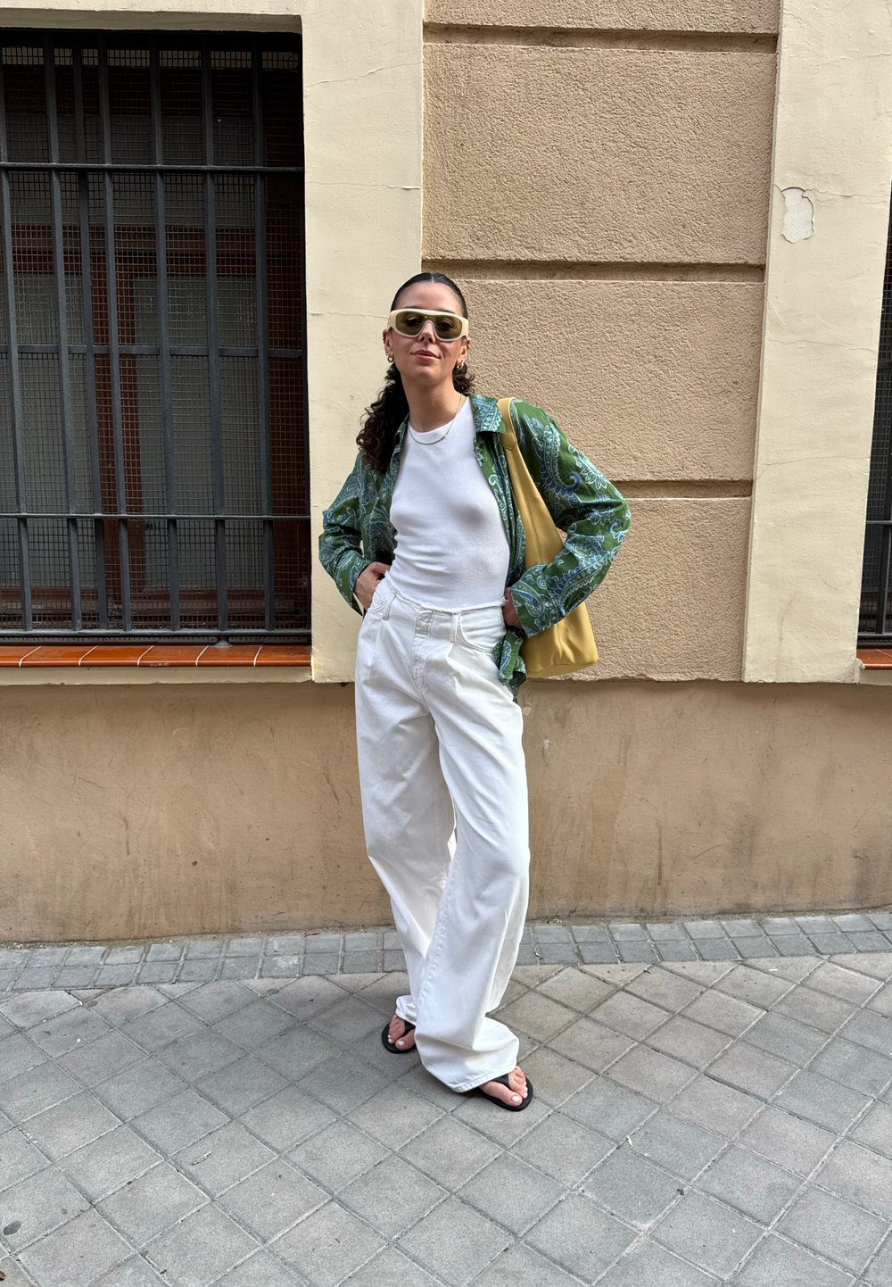 Camisa verde com padrão paisley, top fitted branco, calças largas brancas, bolsa tote amarela e chinelos pretos; estilizado contra uma parede texturizada.