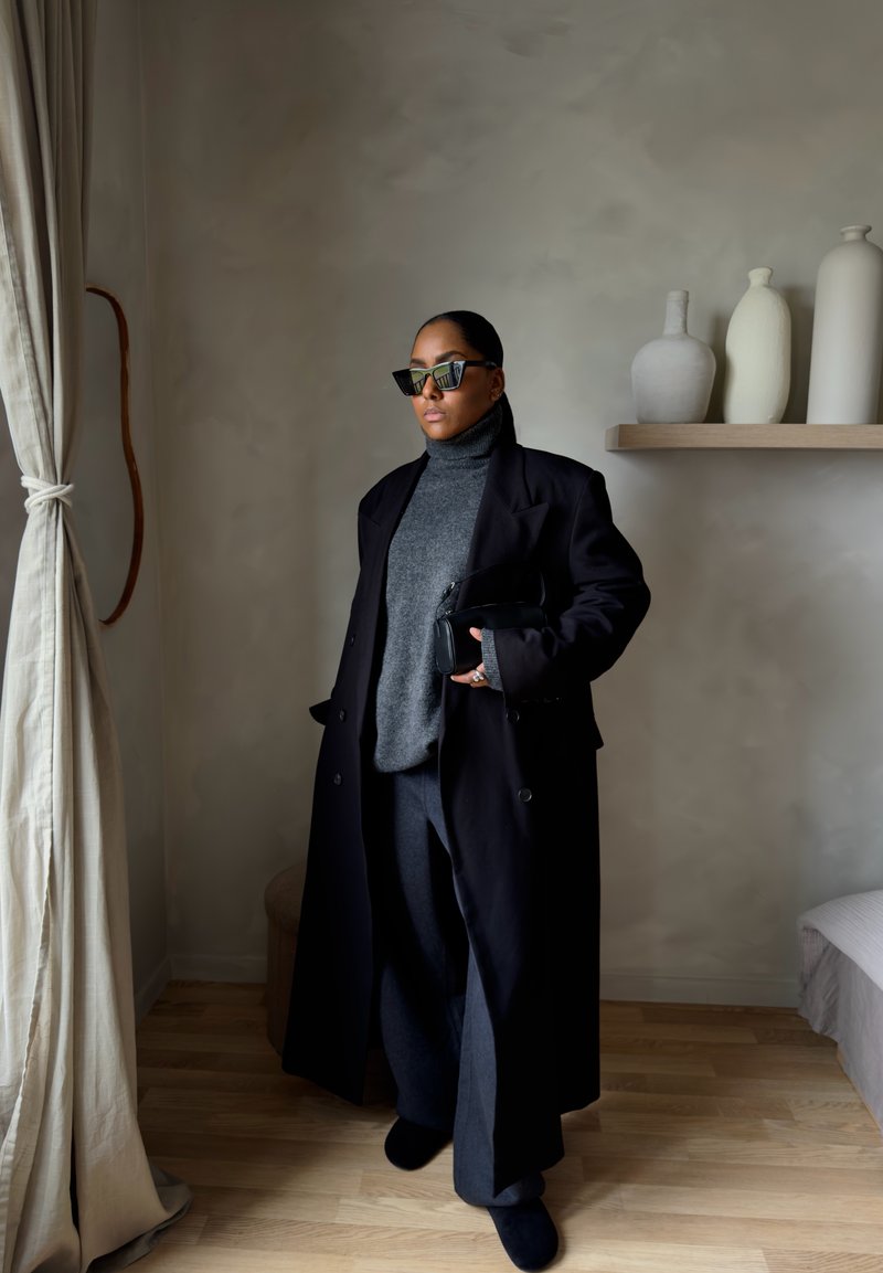 Femme portant un long manteau noir, un pull à col roulé gris, des lunettes de soleil et tenant une pochette noire dans une pièce minimaliste avec des vases sur une étagère.