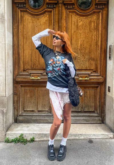 Mujer de pelo rojo con gafas de sol, camiseta negra gráfica de varias capas y falda de encaje blanca, posando frente a una gran puerta de madera al aire libre.
