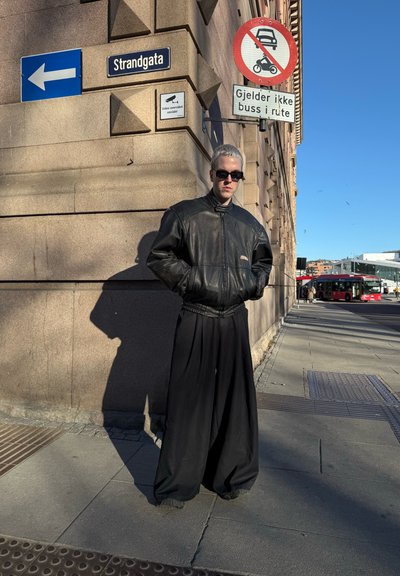 Chaqueta bomber de cuero negro, pantalones negros holgados, de pie contra una pared de piedra. Cielo azul con señales de tráfico en el fondo.