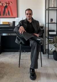 Veste en cuir noire sur un polo noir, jean gris et chaussures noires à semelles épaisses. Style épuré avec des éléments de design minimalistes.