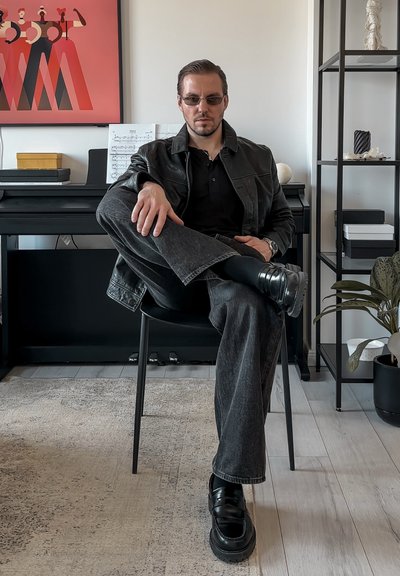 Chaqueta de cuero negra sobre un polo negro, vaqueros grises y zapatos negros con suelas gruesas. Estilo elegante con elementos de diseño minimalista.