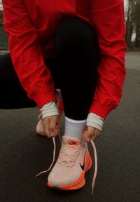 Personne en veste rouge accroupie à l'extérieur, nouant les lacets de chaussures de course Nike roses et orange sur des chaussettes blanches.