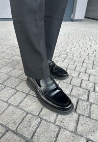 Person som står utendørs på grå brostein iført svarte pen-sko og svarte bukser.