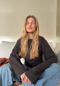 Haut noir à manches longues avec larges poignets, associé à un jean bleu clair taille haute. Cheveux longs et raides encadrant le visage dans une lumière douce.